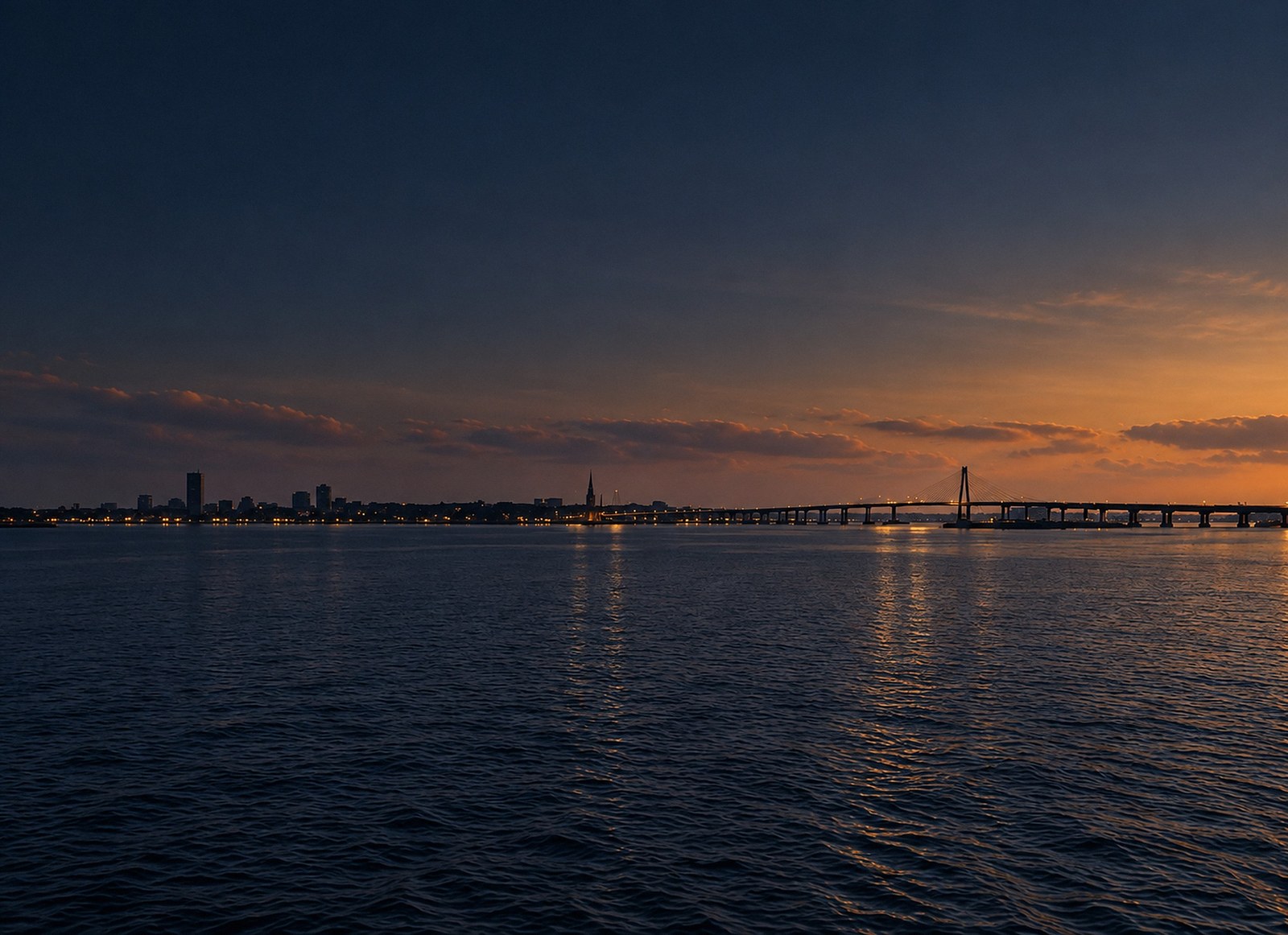 Charleston harbour at dusk, the Ravenel Bridge and the peninsula skyline in silhouette against a low amber horizon.
